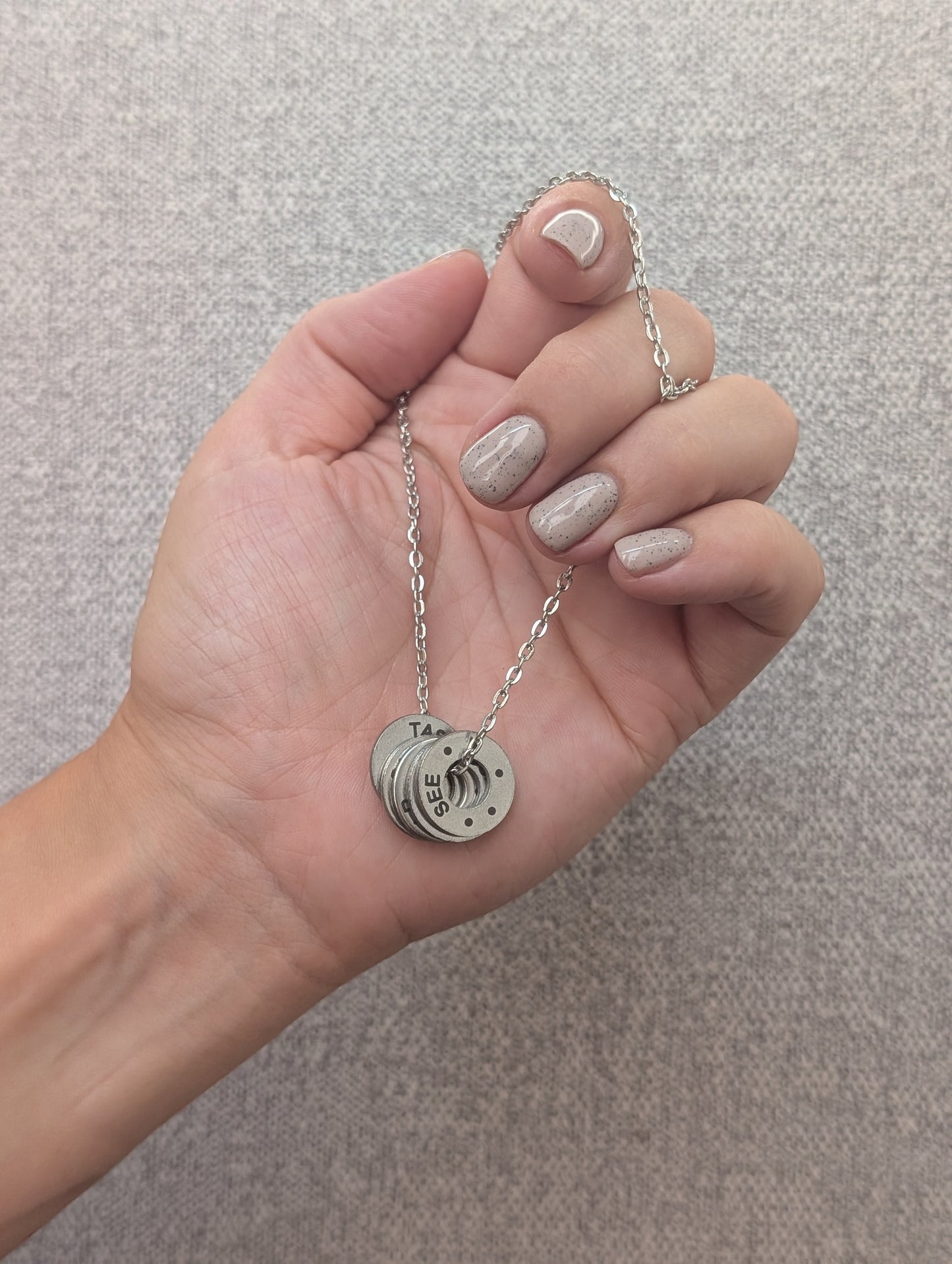 Hand holding a silver necklace with a pendant against a neutral background. Necklace has 54321 grounding technique printed on the pendant.